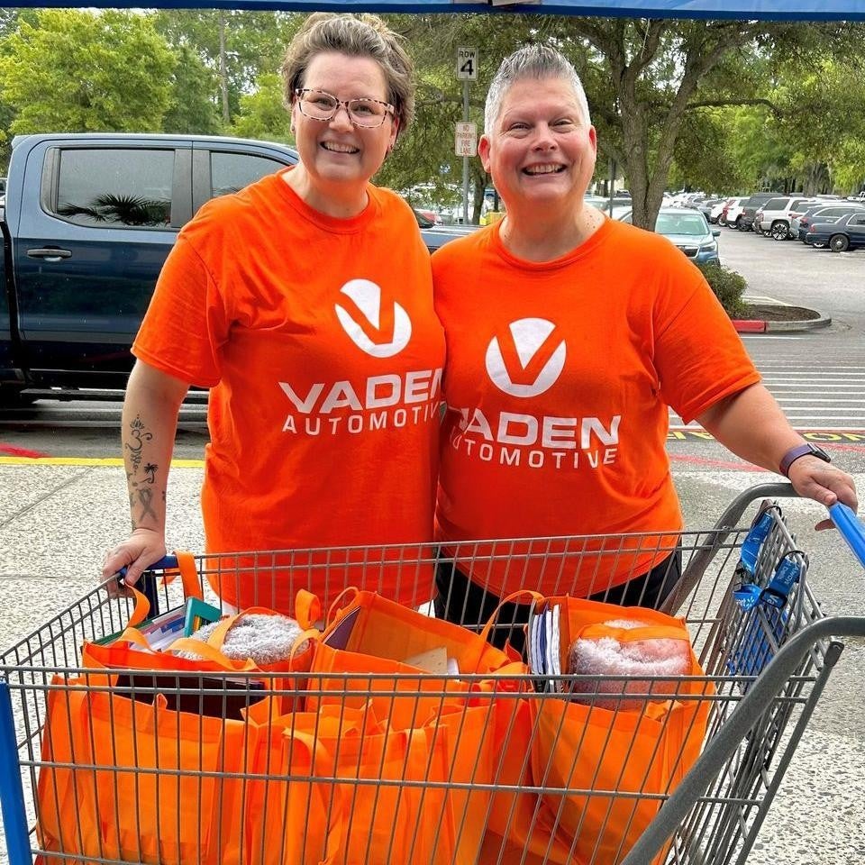 Two people in bright orange shirts that say Vaden Automotive in front of a cart of bright orange bags.